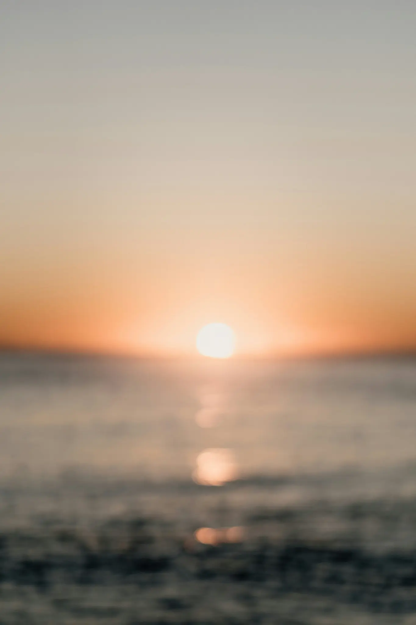 Una foto vertical borrosa de un sol naranja brillante poniéndose sobre agua tranquila y oscura, con un reflejo reluciente.