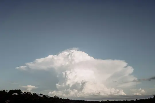 A massive, bright white cumulonimbus cloud with a flat top stretches across a pale blue sky above a dark, tree-covered…
