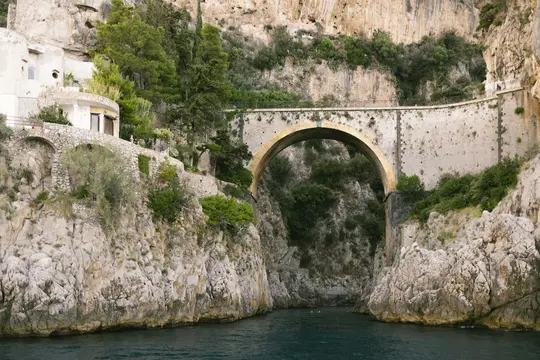 Un ponte ad arco in pietra con un arco giallo-marrone attraversa uno stretto specchio d'acqua blu-verde scuro tra ripide e