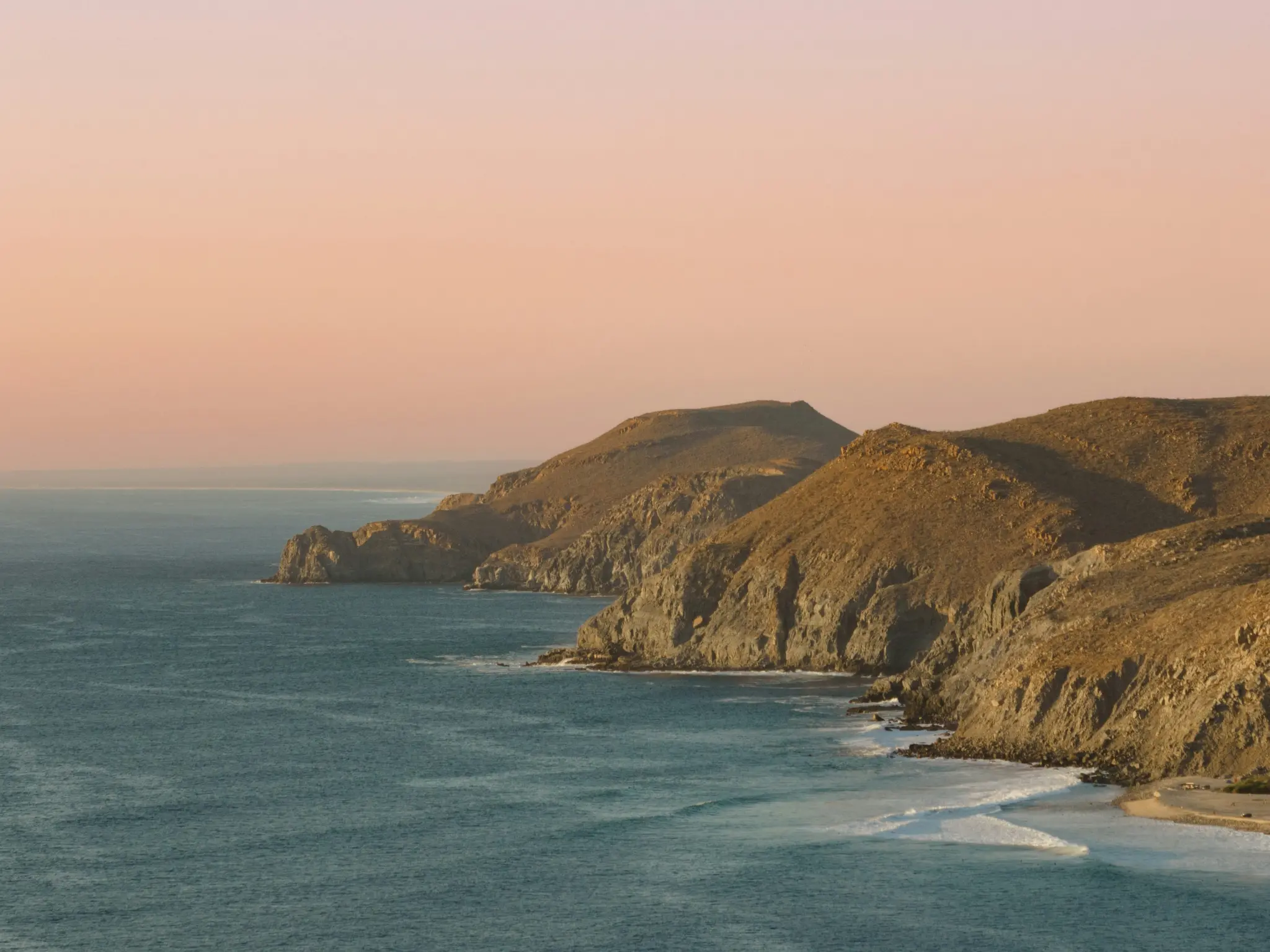 Amplia fotografía paisajística de una escarpada costa al atardecer. Colinas marrones y ocres se encuentran con el océano