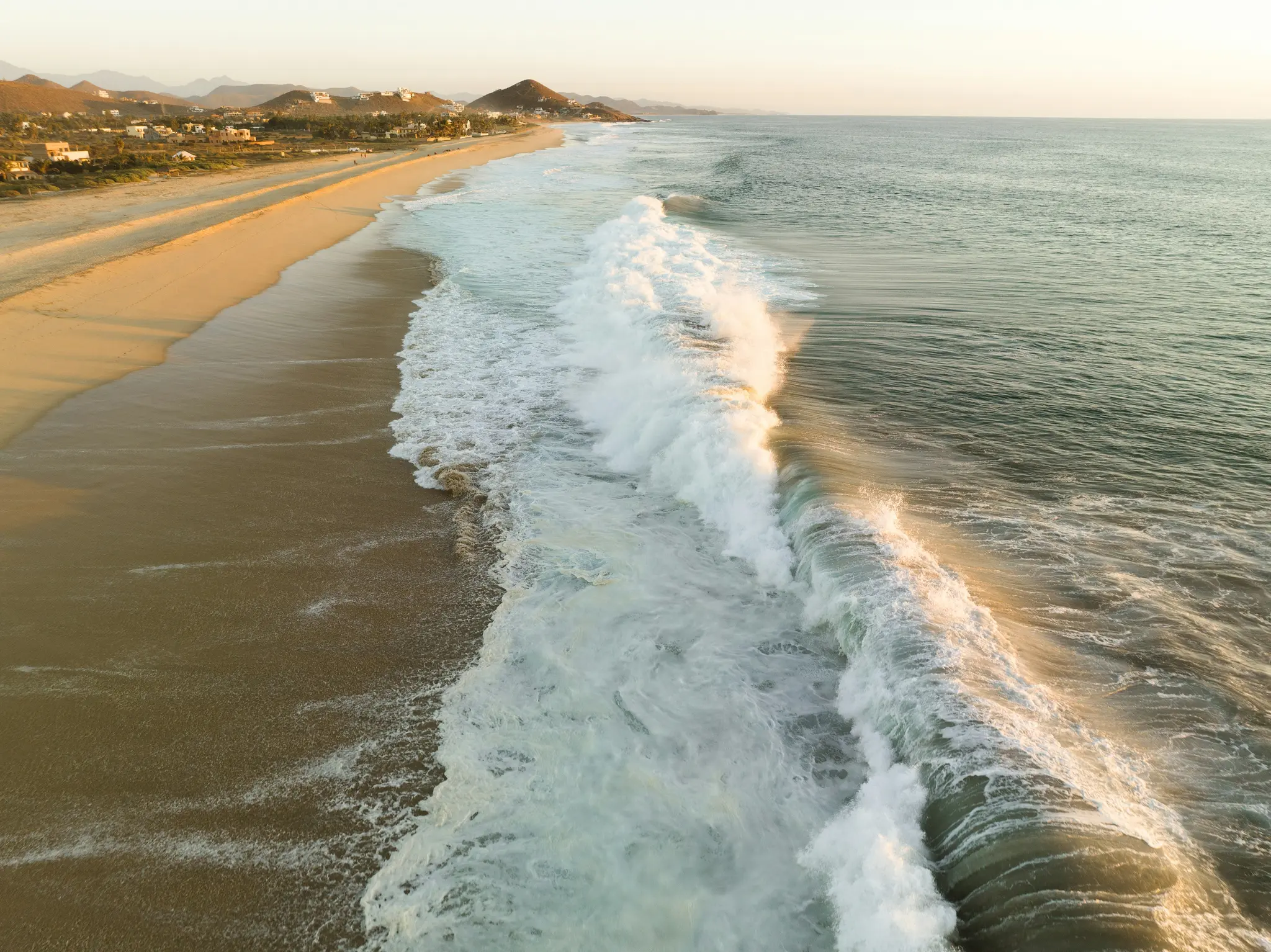 Una vista aérea de una amplia playa de arena a la hora dorada, con grandes olas con crestas blancas que llegan desde el