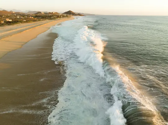 Una vista aerea di un'ampia spiaggia sabbiosa all'ora d'oro, con grandi onde dalle creste bianche che arrivano dall'oceano.