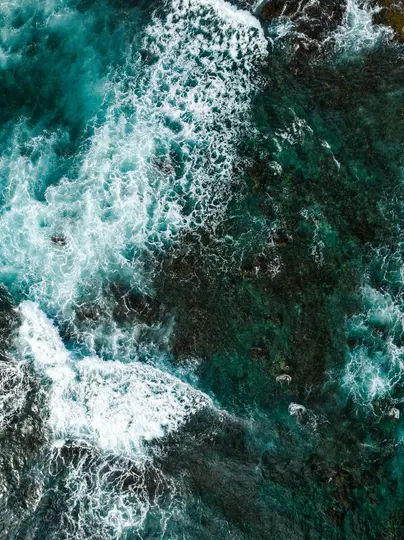 Una vista aerea, dall'alto, mostra onde oceaniche bianche e spumeggianti che si infrangono su rocce scure e sommerse in acqua
