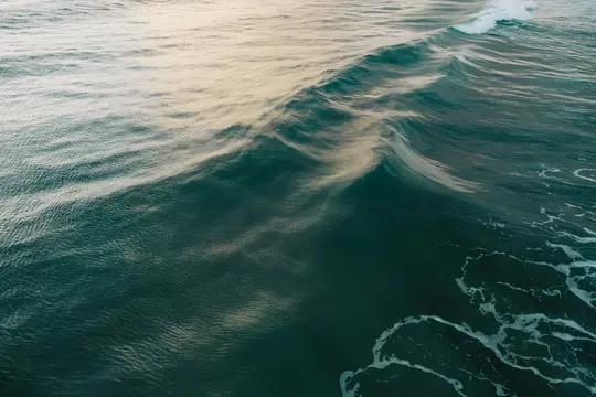 Un primo piano aereo di onde oceaniche. L'acqua è di un blu turchese intenso, con un'onda prominente che si infrange al
