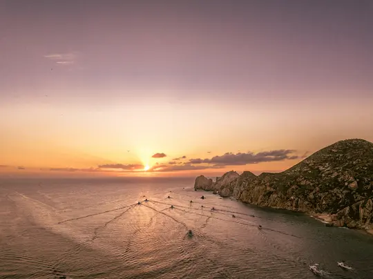 Eine Luftaufnahme einer lebendigen Sonnenuntergangslandschaft über dem Meer mit zahlreichen Booten, die V-förmige Kielwasser