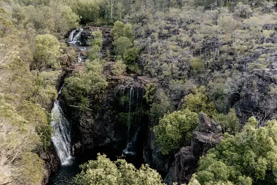 Eine Luftaufnahme einer Reihe von Wasserfällen, die über dunkle, felsige Klippen mit üppigem Grün und hellen Bäumen