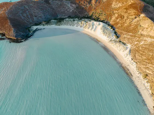 Eine Luftaufnahme einer Bucht mit klarem türkisfarbenem Wasser, einem sandigen, mondsichelförmigen Strand und trockenen