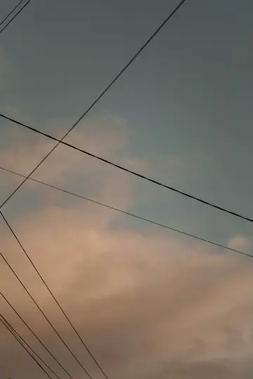 Ein Himmel mit weichen, blassblauen und orangefarbenen Wolken, durchzogen von mehreren dunklen, dünnen Stromleitungen, die