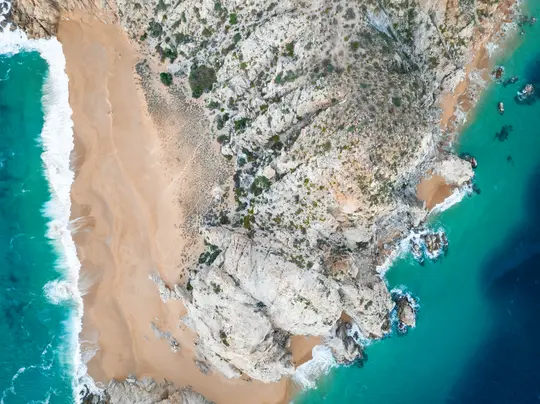 Eine Luftaufnahme, die direkt nach unten auf einen Sandstrand, Felsklippen und das Meer mit türkisfarbenem und tiefblauem