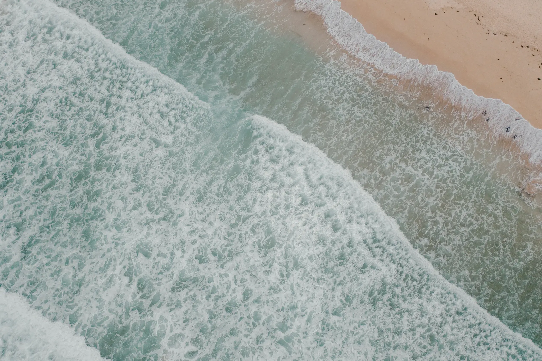 Et luftfoto viser lyseblå havbølger med hvidt skum, der bryder på en beige sandstrand. Vandet går fra…
