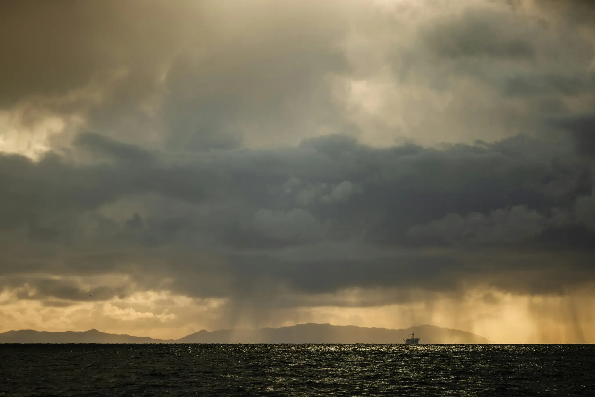 A small boat floats on dark, choppy water under a dramatic, cloudy sky with shafts of rain visible in the distance.