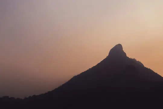 Ein dunkler, zerklüfteter Berggipfel hebt sich gegen einen Himmel ab, der vom hellen Orange am Horizont zu einem sanften Rosa