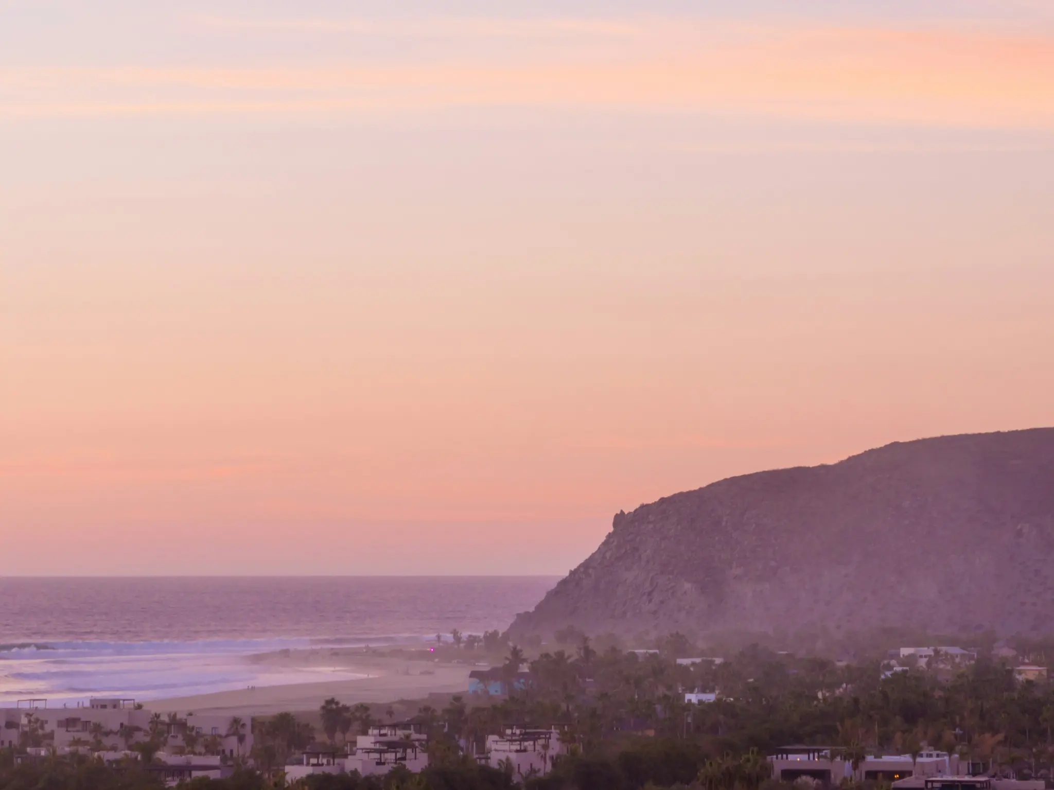 An aerial view of a beach and ocean at sunset, with a large hill on the right and buildings and palm trees below.