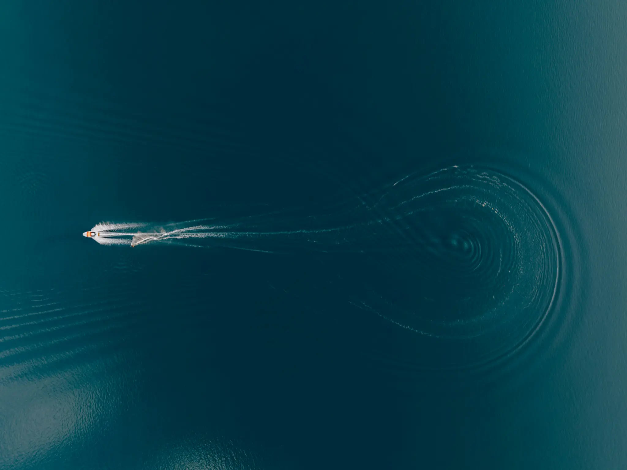 An aerial shot of a small white and orange boat moving left across dark blue water, leaving a long white wake and circular…
