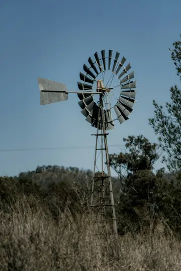 Eine verwitterte Metallwindmühle mit großem Ventilator und Leitwerk steht vor einem klaren blauen Himmel, umgeben von