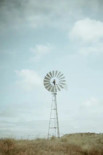 A tall, weathered metal windmill with a circular fan stands in a field of dry, light brown and green grass under a pale…