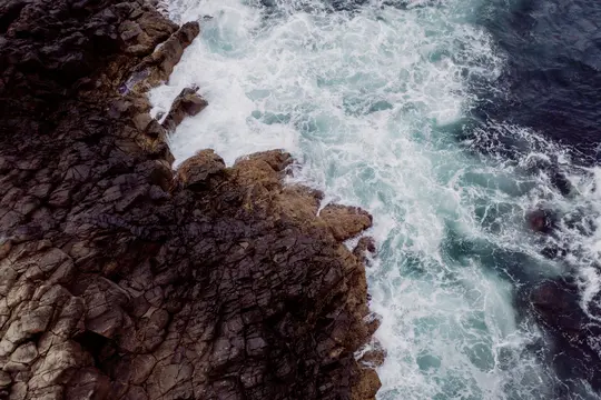 Una vista aerea mostra rocce scure e texturizzate a sinistra, che incontrano l'acqua dell'oceano schiumosa, bianca e turchese