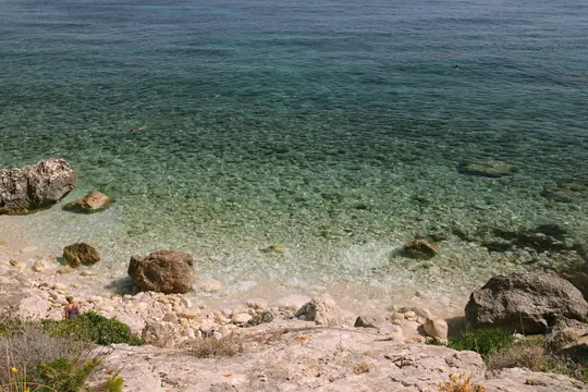 Eine Ansicht von oben auf ein klares türkisfarbenes Meer, das auf eine felsige und kieselige Küste trifft. Ein Schwimmer ist