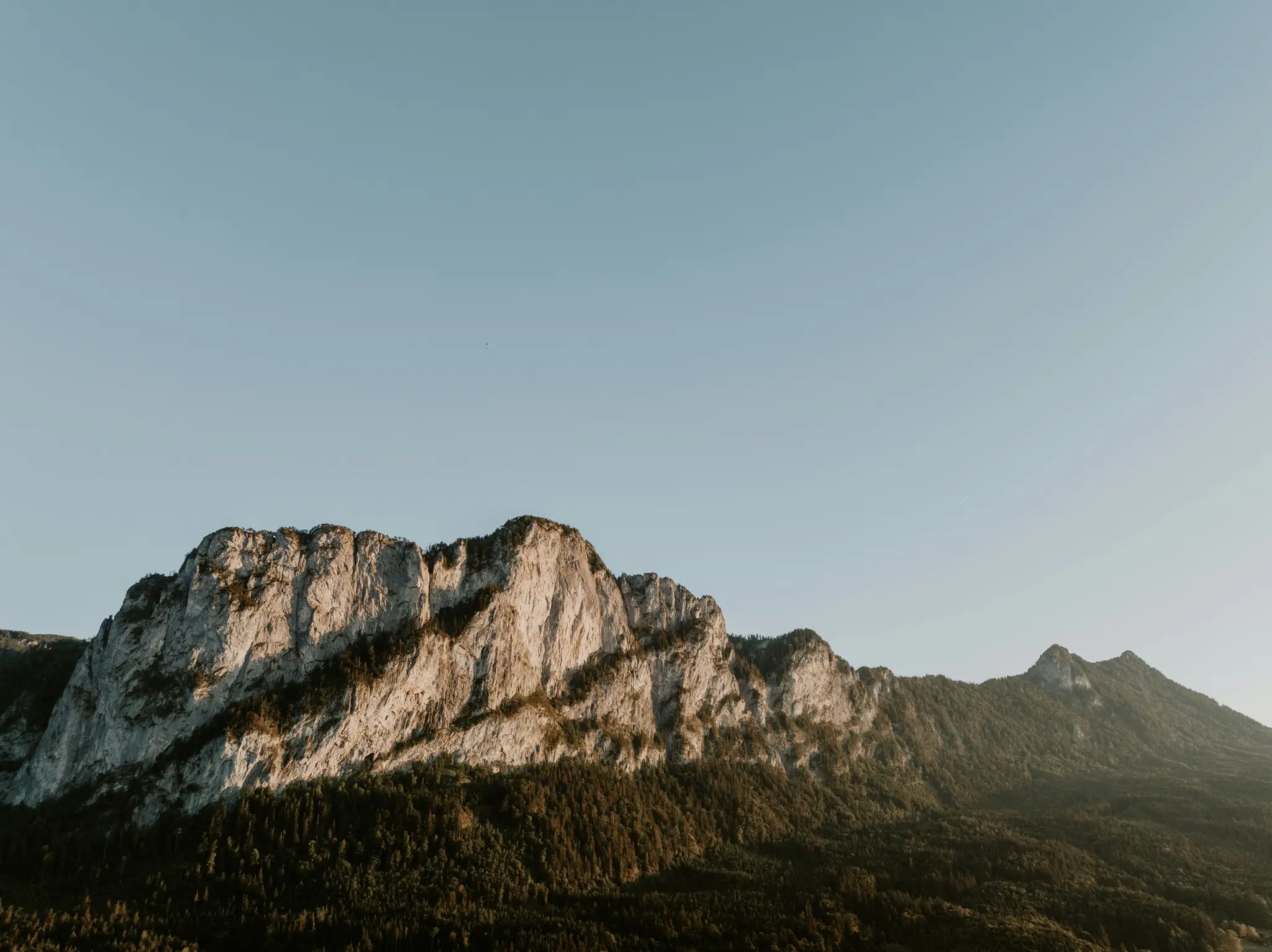 Ett brett landskapsfoto som visar en bergskedja med robusta, ljusa klippor och mörkgröna skogar nedanför. Himlen är...