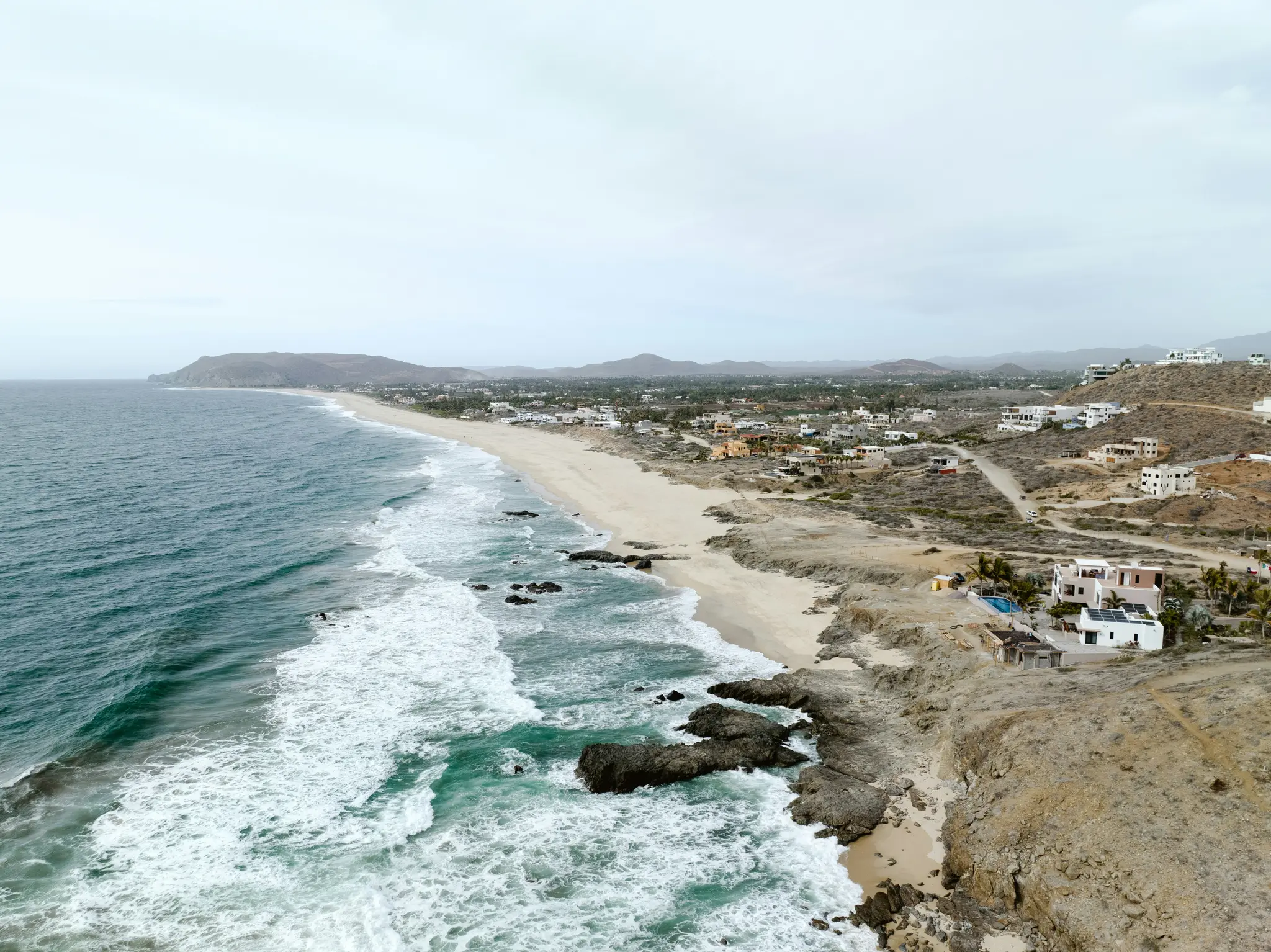Et luftfoto viser en lang sandstrand med turkisblå havbølger, der bryder mod kysten og mørke klipper. En by...