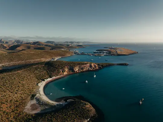 Una foto aerea mostra una baia curva con acqua turchese, diverse barche e una spiaggia sabbiosa. Si ergono colline aride