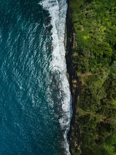 An aerial shot shows the deep blue ocean on the left, with white waves crashing against a dark, rocky cliff. The right side…