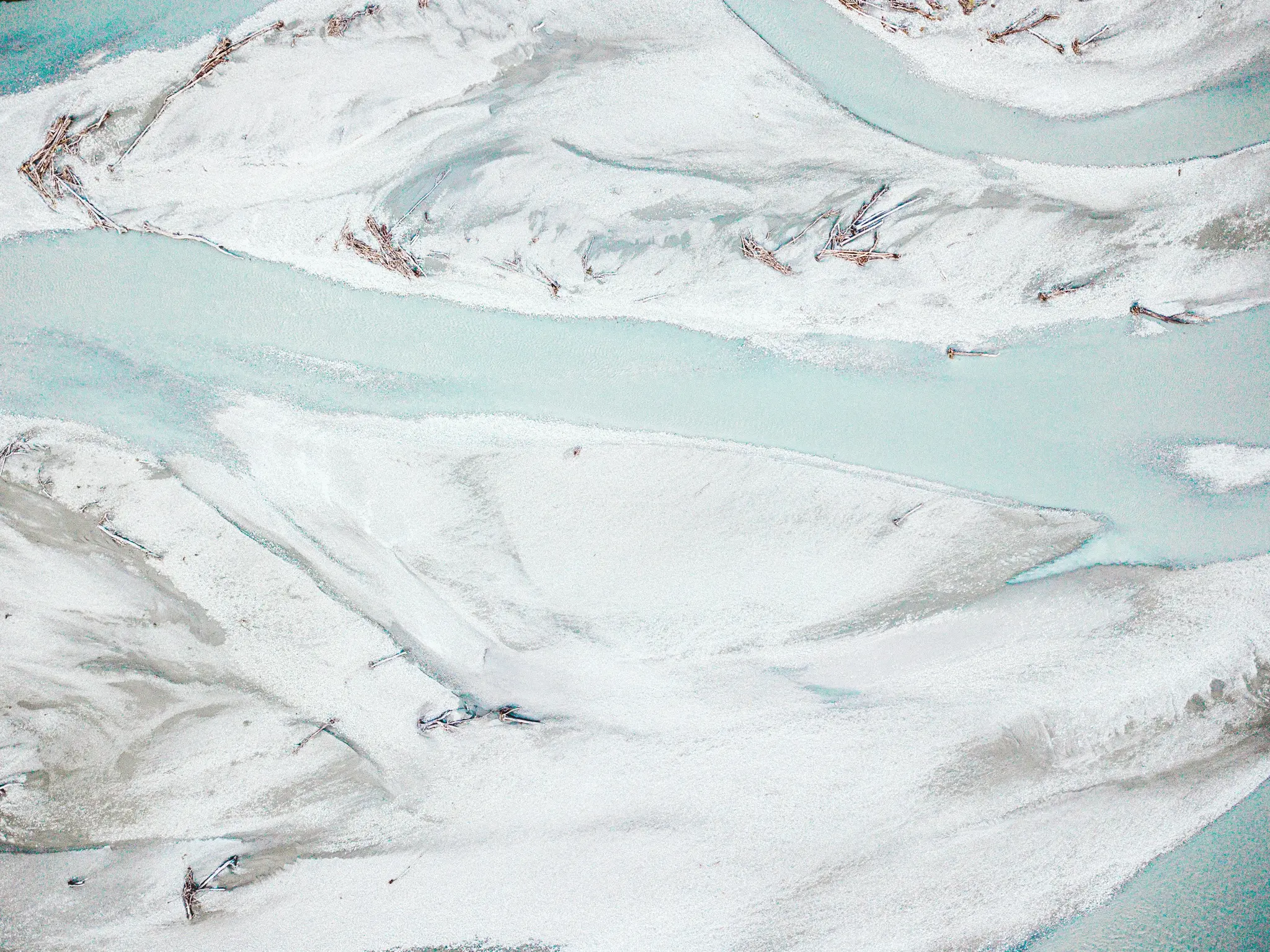 An aerial shot shows a light blue river winding through white and light grey sandbars, forming a braided pattern. Driftwood…
