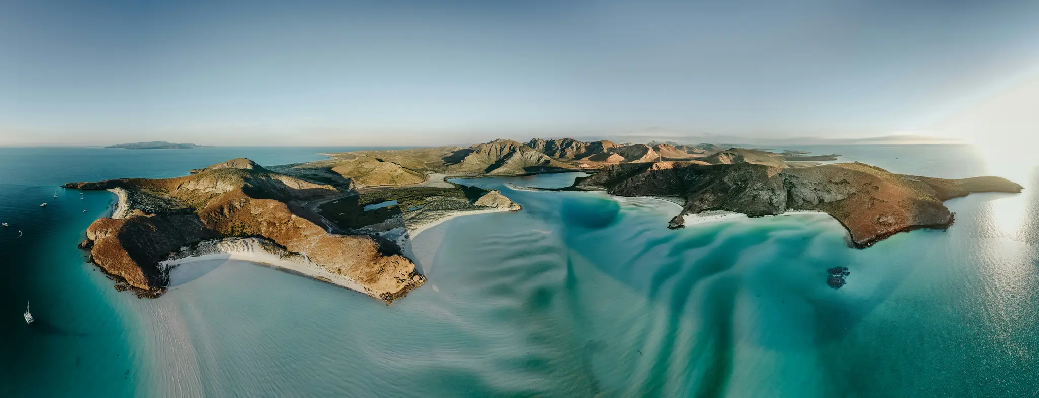 En luftbildad panoramavy över en karg kustlinje med flera öar med bruna och gröna berg, vit sand…