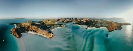 En luftbildad panoramavy över en karg kustlinje med flera öar med bruna och gröna berg, vit sand…