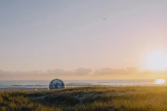 A vibrant sunrise over a beach, with a blue, semi-circular art installation on a grassy dune in the foreground.