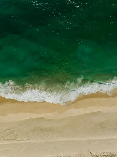 Una vista aerea, dall'alto, di una spiaggia sabbiosa color oro che incontra l'oceano verde intenso e turchese. Onde bianche…