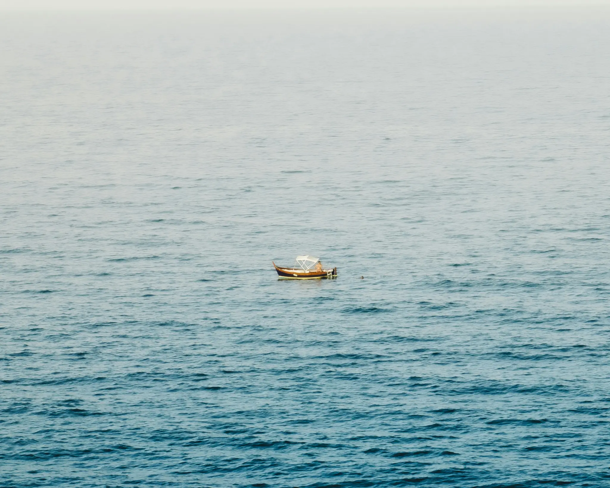 A small, light brown wooden boat with a white canopy floats alone on a vast expanse of calm, rippling blue water under a…
