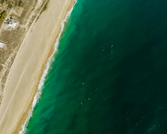 An aerial shot of a long, light-beige sandy beach on the left, meeting a vibrant turquoise and deep green ocean on the…