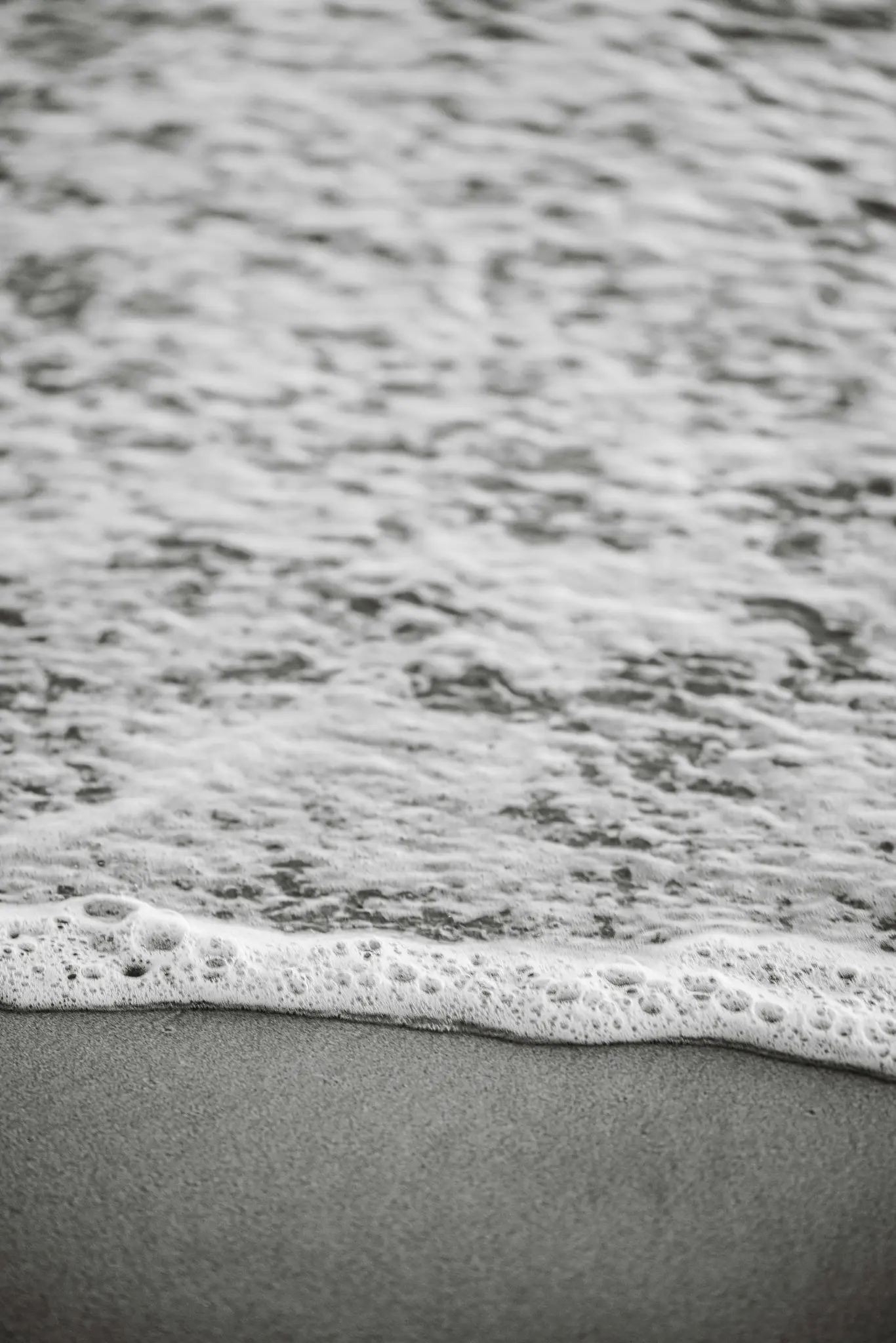 Schwarz-Weiß-Nahaufnahme von schaumigen Meereswellen, die sanft über einen glatten, dunklen Sandstrand spülen.