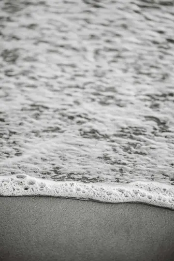 Schwarz-Weiß-Nahaufnahme von schaumigen Meereswellen, die sanft über einen glatten, dunklen Sandstrand spülen.