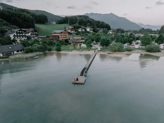 Eine Luftaufnahme zeigt einen langen Holzsteg, der in einen ruhigen See ragt. Ein Dorf mit Häusern und einem Campingplatz