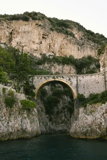A stone arch bridge with a golden-hued underside spans a dark blue inlet between towering, vegetation-covered cliffs.