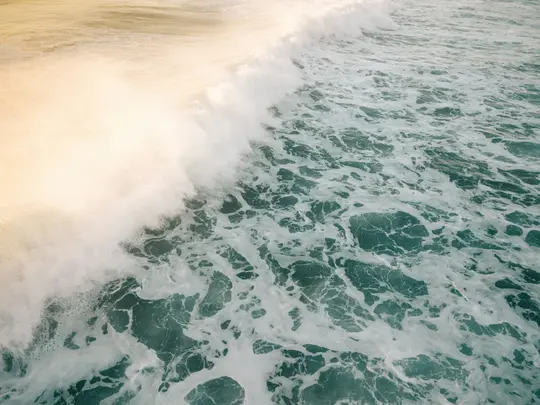 A high-angle shot of an ocean wave breaking, showing a large mass of white foam against dark teal water.