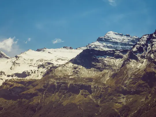 Breitformatige Landschaftsaufnahme einer Bergkette mit schneebedeckten Gipfeln und felsigen, bewachsenen unteren Hängen unter
