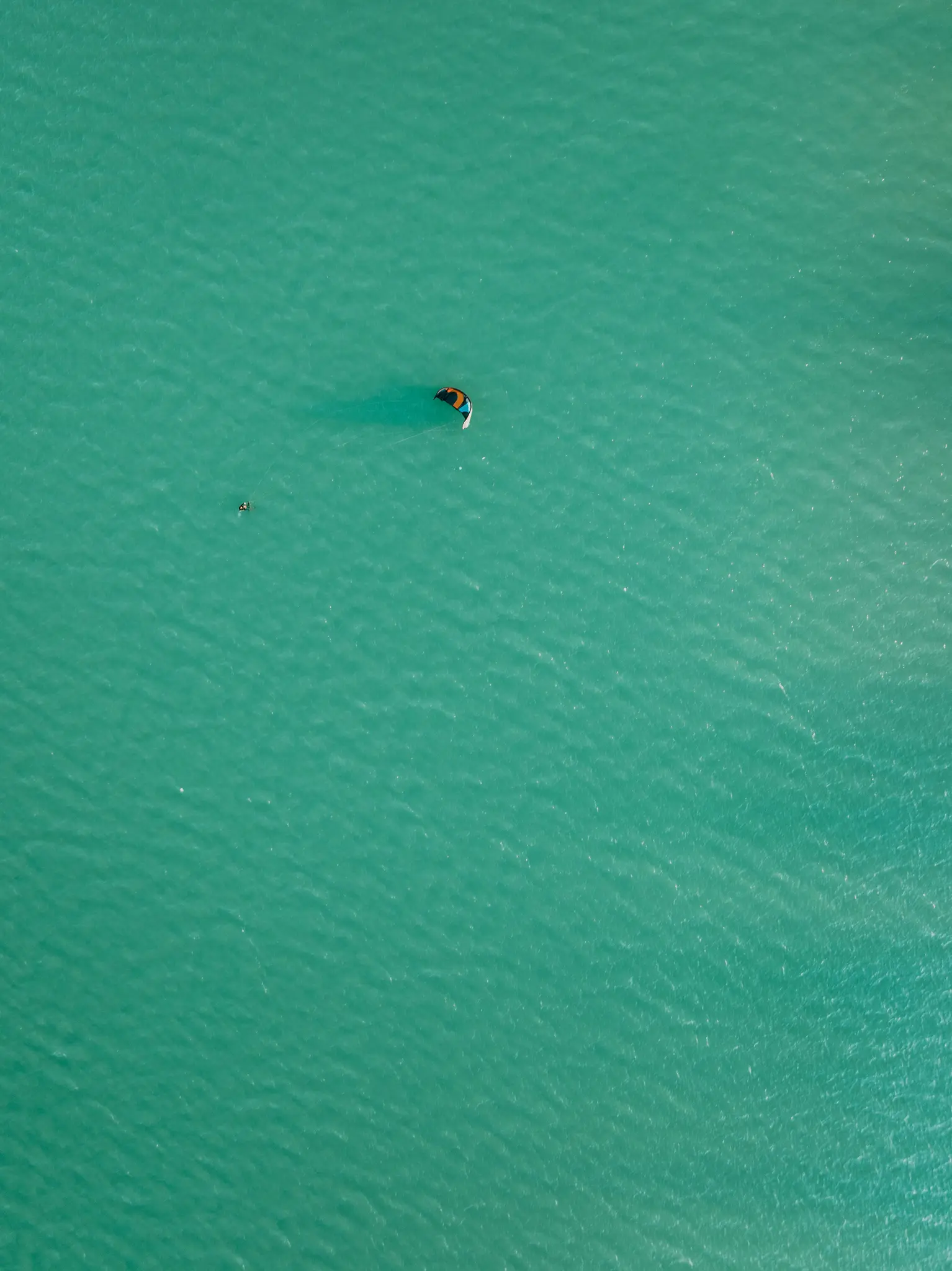 En flygvy över en ensam kitesurfare på ett vidsträckt, skimrande turkost vatten under starkt dagsljus.