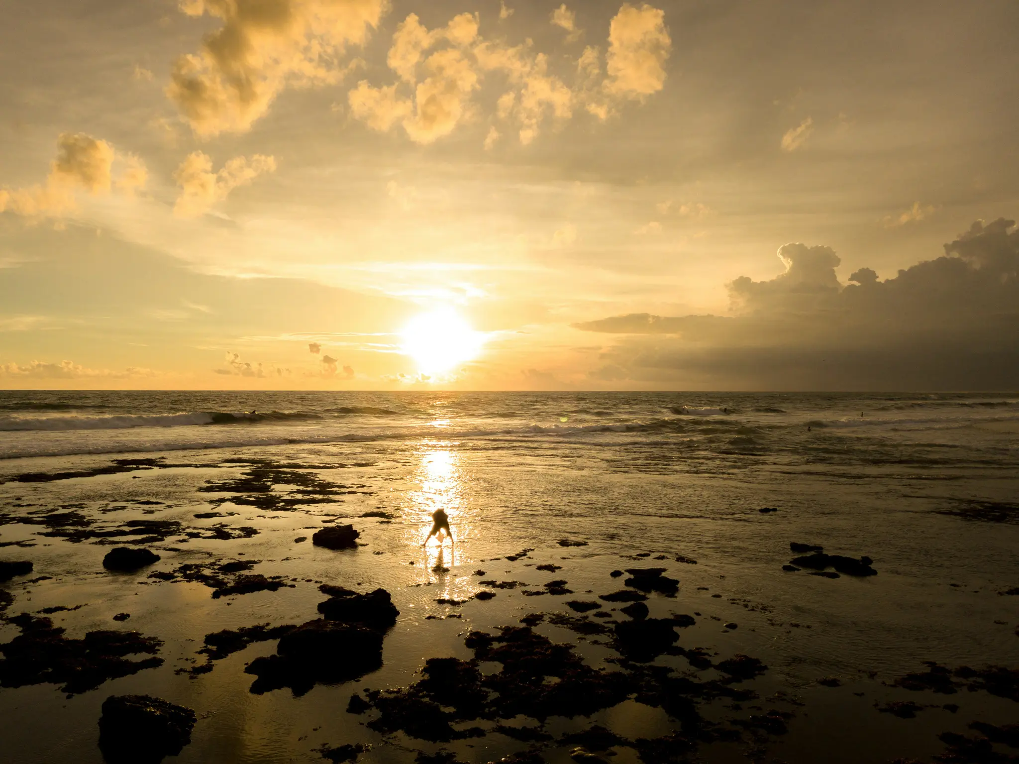 A vibrant sunset over a rocky ocean shore. The sun, a bright white orb, reflects on the wet sand and water. A lone figure…