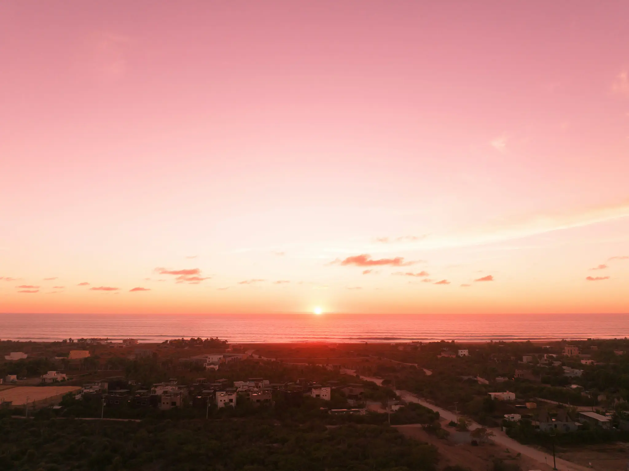 Een luchtfoto van een zonsondergang boven een kustplaats. De zon is een heldere bol aan de horizon en werpt roze en oranje