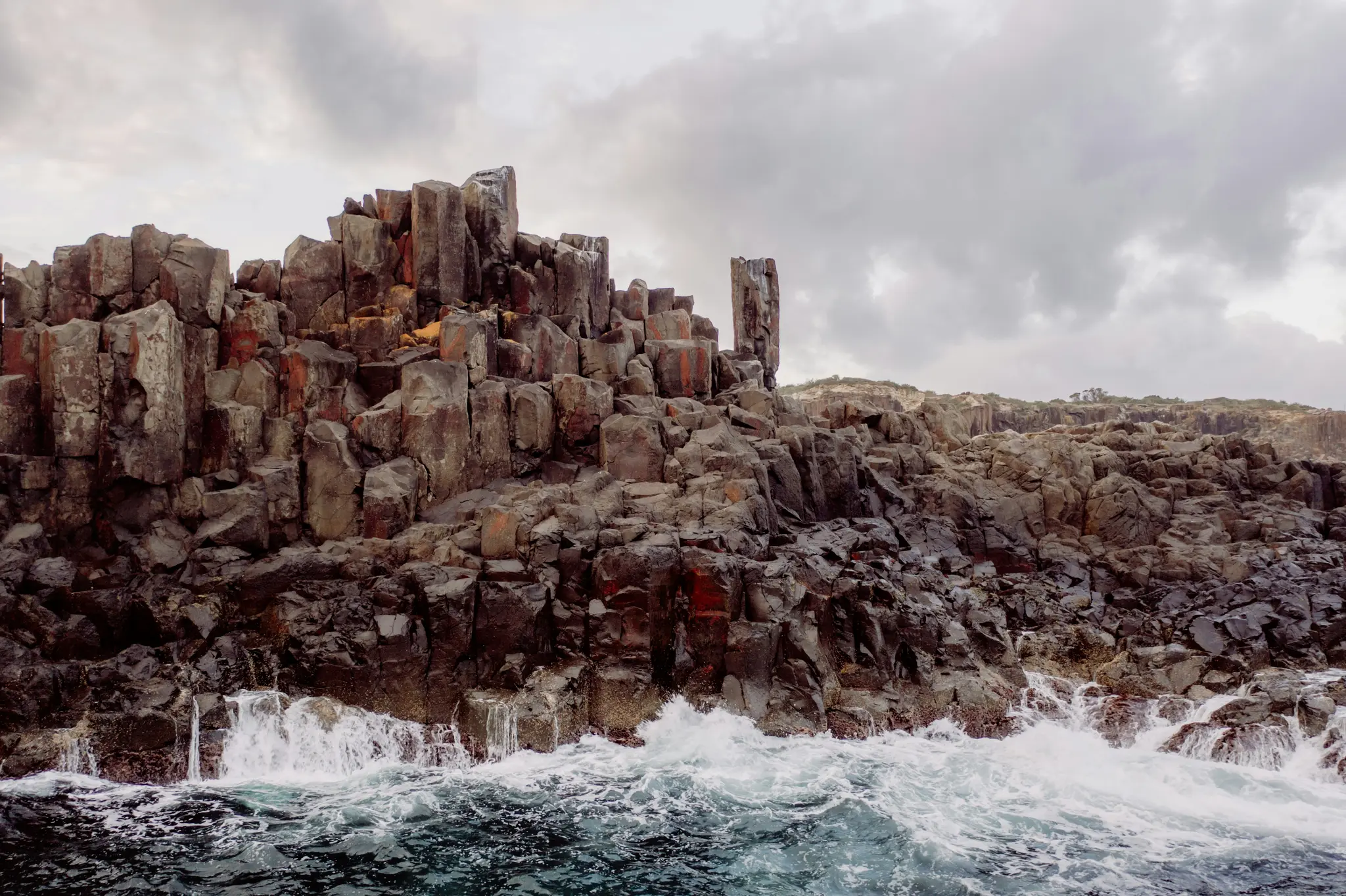 Una toma panorámica de acantilados de basalto columnar oscuros y angulosos que se elevan desde el océano. Olas blancas rompen