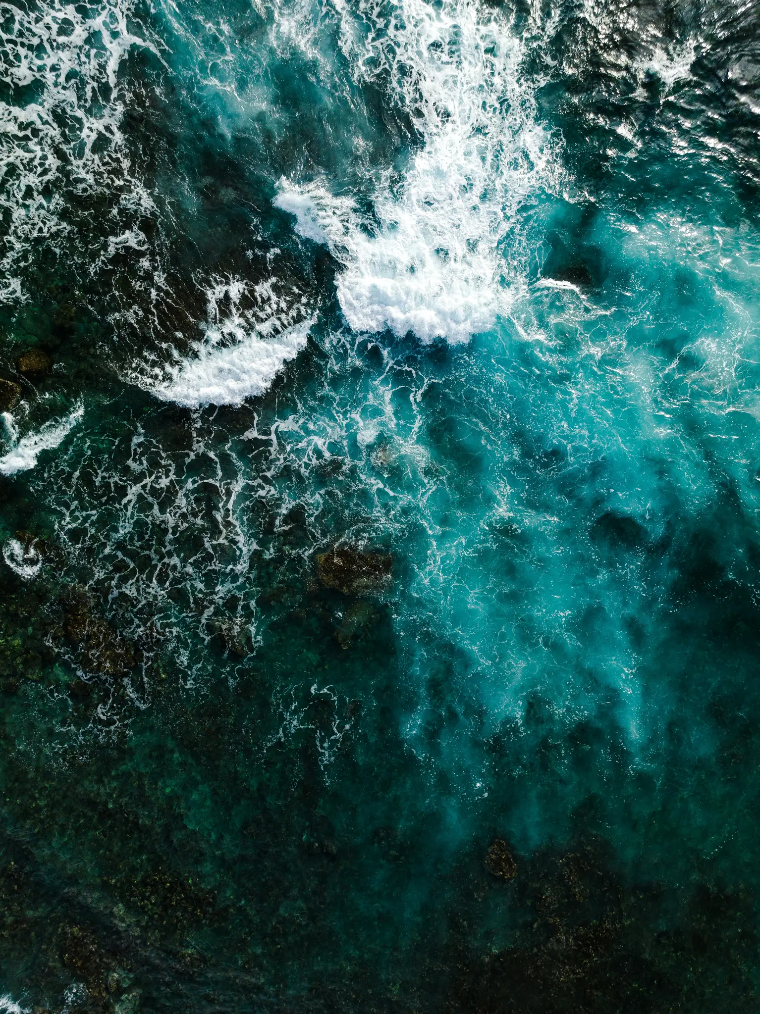 An aerial shot looking down at the ocean, showing white waves crashing over dark rocks and revealing clear blue and green…