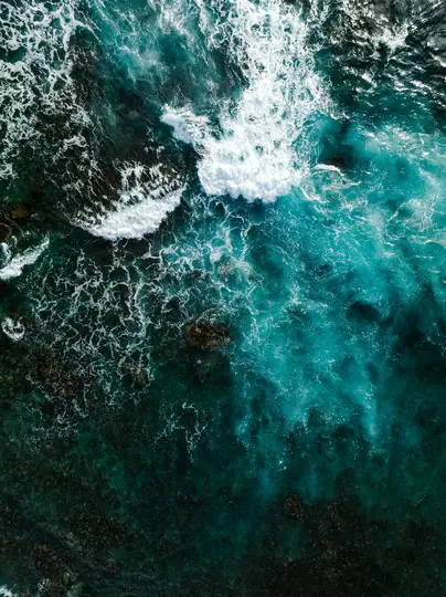 An aerial shot looking down at the ocean, showing white waves crashing over dark rocks and revealing clear blue and green…