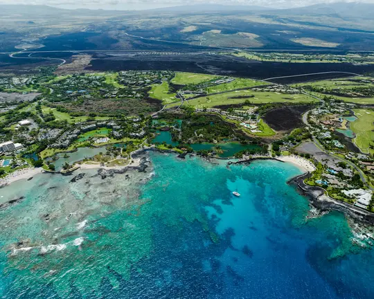 An aerial photo captures a vibrant coastal resort with green golf courses, buildings, and lagoons beside turquoise ocean…