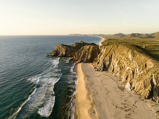 Eine Luftaufnahme zeigt einen langen Sandstrand, der rechts von rauen, felsigen Klippen und dem blauen Ozean mit weißen...