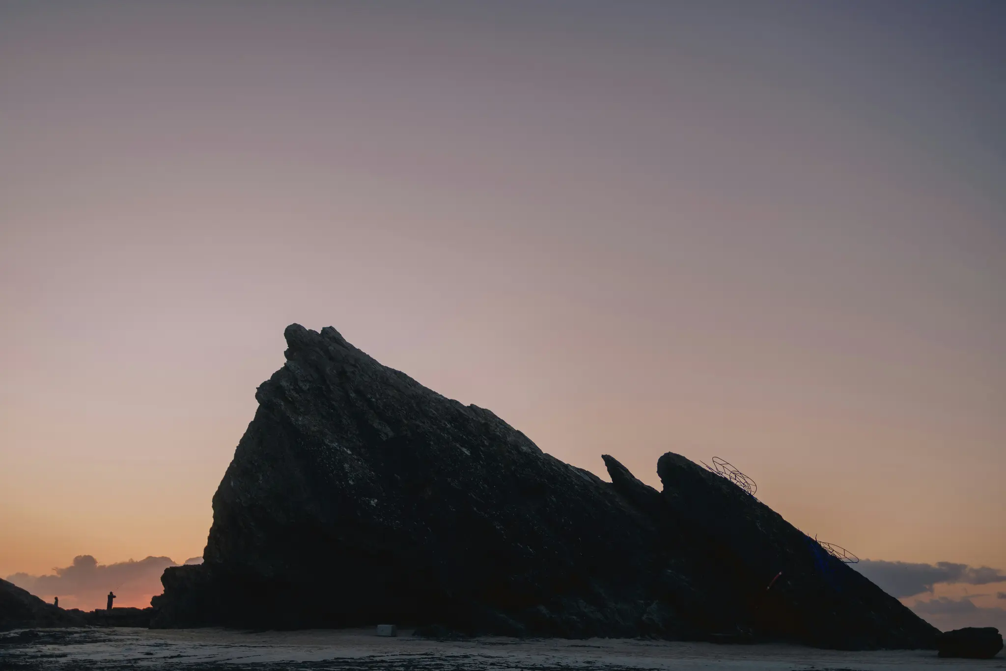 A large, dark, jagged rock formation is silhouetted against a sky transitioning from orange at the horizon to purple and…