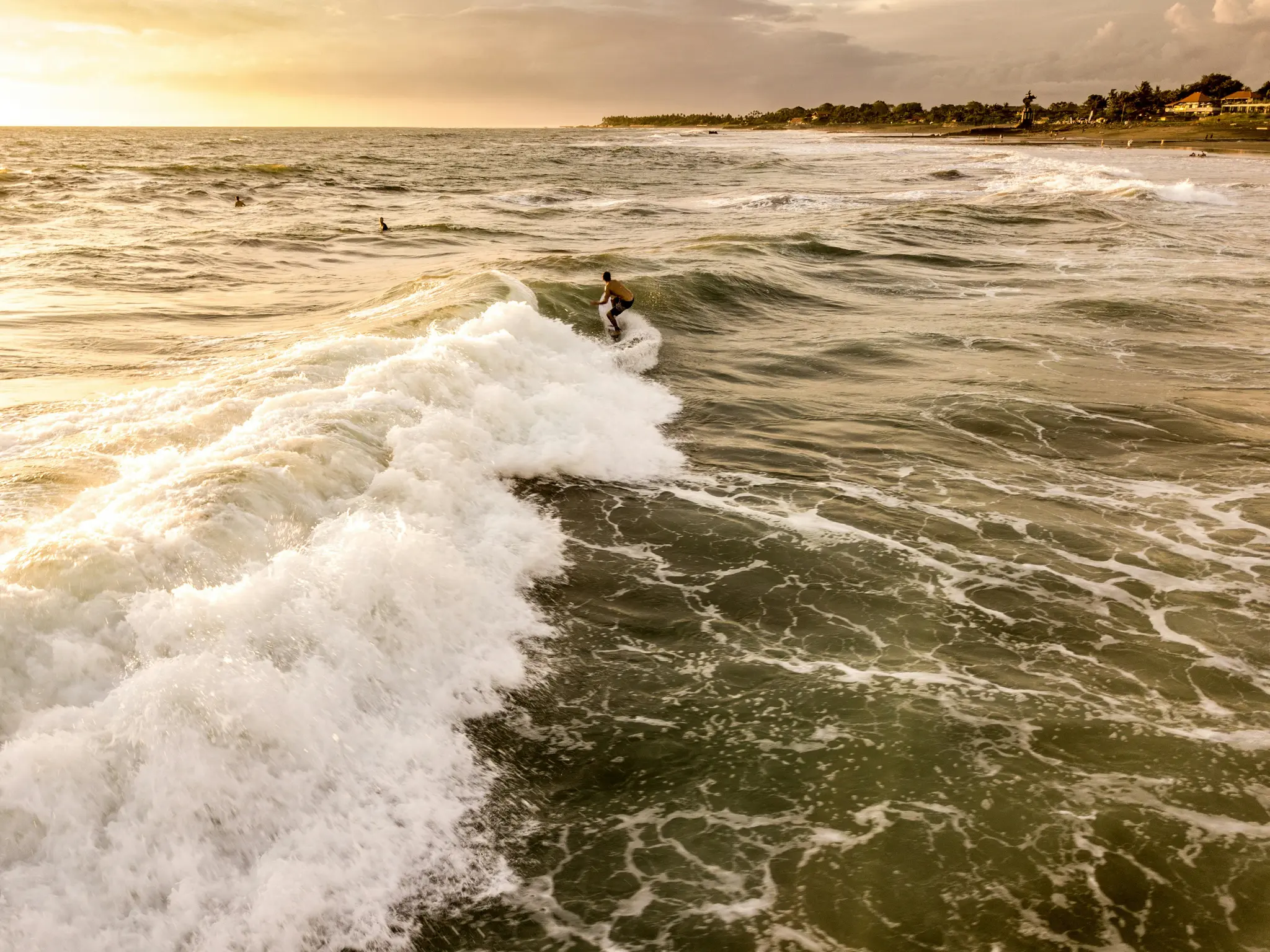 A surfer in a yellow shirt rides a large white-foamed wave on a golden ocean. The sun sets, casting a warm glow.