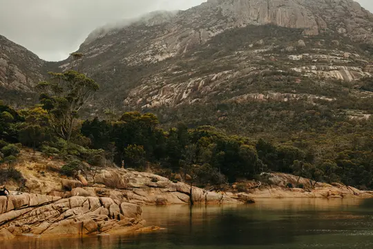 A wide landscape photo shows a calm body of water in the foreground, leading to a rocky shore with green trees. A large…